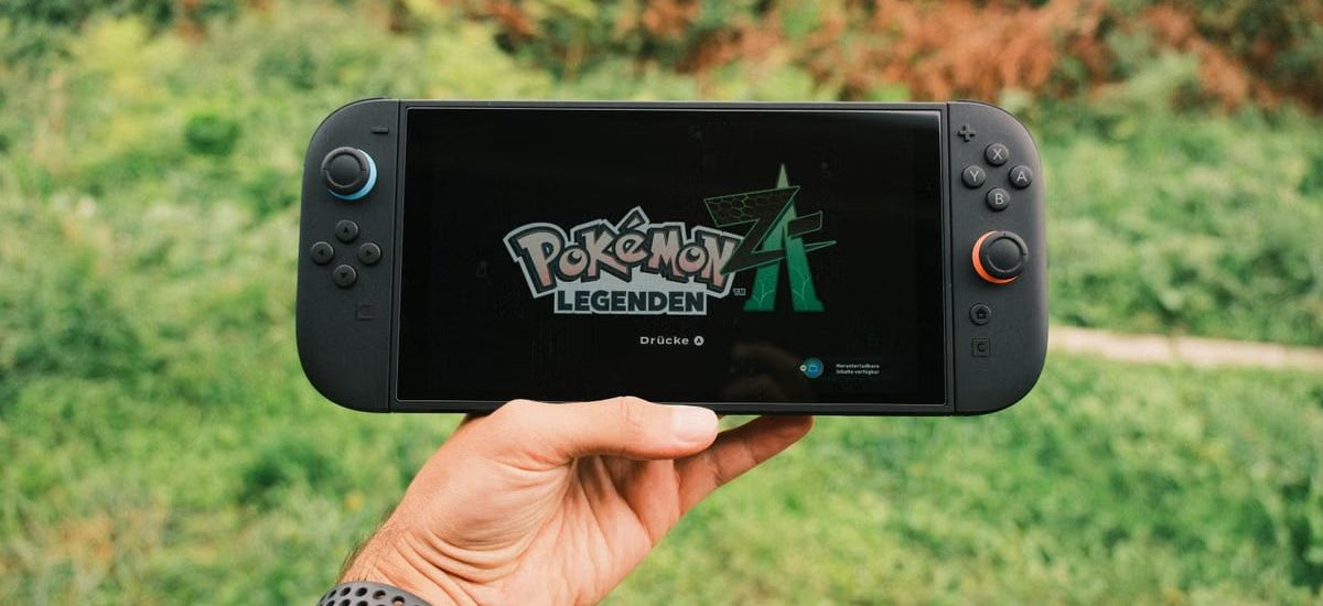 A hand holding a handheld gaming console displaying the Pokémon Legends game screen outdoors.
