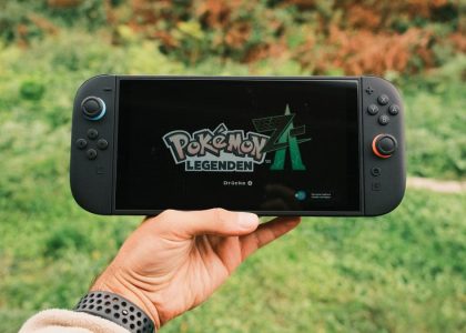 A hand holding a handheld gaming console displaying the Pokémon Legends game screen outdoors.